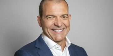 Smiling man in a navy blazer and white shirt against a gray background (professional headshot).