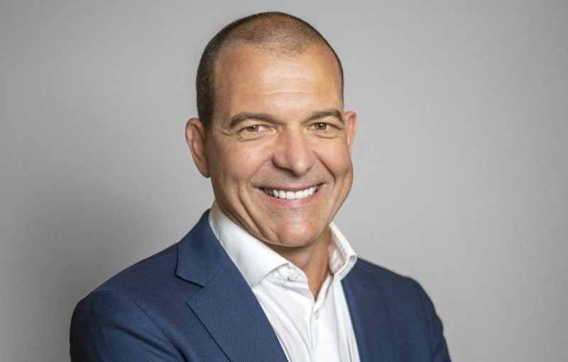 Smiling man in a navy blazer and white shirt against a gray background (professional headshot).