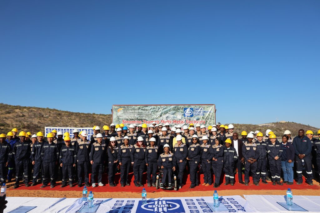 MMR Group Photo | Mining and Energy Namibia | Namibia’s Leading Mining & Energy News Group of workers in dark blue uniforms and yellow/white hard hats posing on a red carpet at a mining site, banner behind reads Sinomine Tsumeb Multi-Metal Recovery