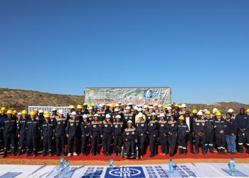 MMR Group Photo | Mining and Energy Namibia | Namibia’s Leading Mining & Energy News Group of workers in dark blue uniforms and yellow/white hard hats posing on a red carpet at a mining site, banner behind reads Sinomine Tsumeb Multi-Metal Recovery