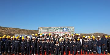 MMR Group Photo | Mining and Energy Namibia | Namibia’s Leading Mining & Energy News Group of workers in dark blue uniforms and yellow/white hard hats posing on a red carpet at a mining site, banner behind reads Sinomine Tsumeb Multi-Metal Recovery