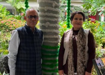 Two adults pose in a lush garden, standing beside a tree wrapped with green and white fabric in the background.