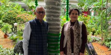 Two adults pose in a lush garden, standing beside a tree wrapped with green and white fabric in the background.