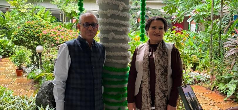 Two adults pose in a lush garden, standing beside a tree wrapped with green and white fabric in the background.