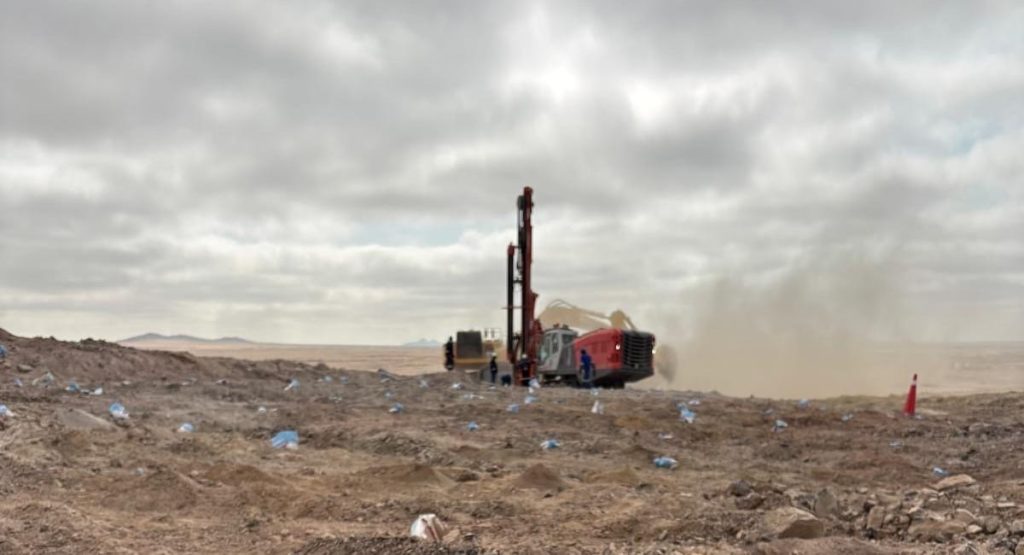Red drilling rig operates in a dusty, barren desert with a few workers nearby under a cloudy sky