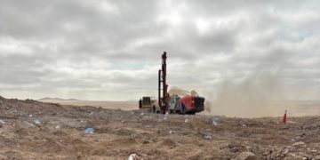 Red drilling rig operates in a dusty, barren desert with a few workers nearby under a cloudy sky