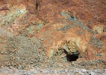 Rugged hillside of orange-brown rock with a dark mine entrance at center-right, surrounded by loose stones and rubble