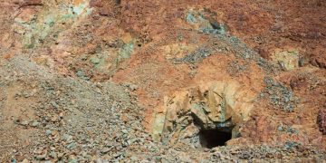 Rugged hillside of orange-brown rock with a dark mine entrance at center-right, surrounded by loose stones and rubble