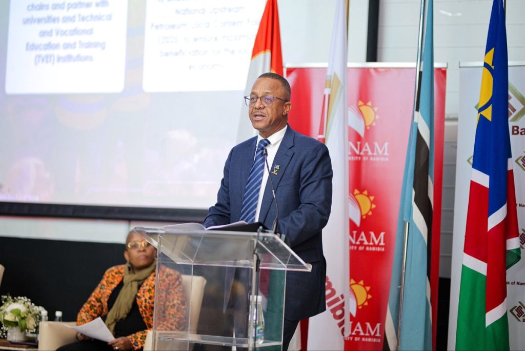 WhatsApp Image 2026-08-16 at 94918 PM | Mining and Energy Namibia | Namibia’s Leading Mining & Energy News Man in a navy suit and glasses speaks at a clear lectern during a formal event, with multiple national flags and NAM banners behind him.