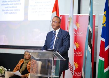 Man in a navy suit and glasses speaks at a clear lectern during a formal event, with multiple national flags and NAM banners behind him.