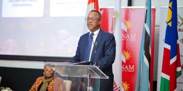 WhatsApp Image 2026-08-16 at 94918 PM | Mining and Energy Namibia | Namibia’s Leading Mining & Energy News Man in a navy suit and glasses speaks at a clear lectern during a formal event, with multiple national flags and NAM banners behind him.