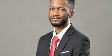Portrait of a man in a black suit with a red tie, looking at the camera against a gray background.