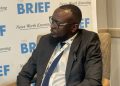 Man in a dark suit and glasses seated in a discussion, with a backdrop that says 'THE BRIEF' and 'News Worth Knowing'.