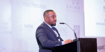 Man in a dark suit speaks at a conference podium with a microphone, facing right.