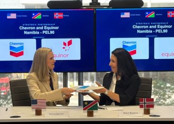 Two women in business attire exchange documents at a conference table, with US, Namibia, and Norway flags, nameplates Beatrice Bienvenu and Aynur Rzayeva, and Chevron/Equinor logos on large blue screens behind.