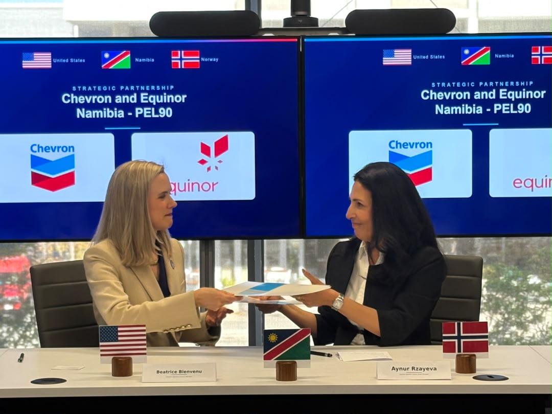 Two women in business attire exchange documents at a conference table, with US, Namibia, and Norway flags, nameplates Beatrice Bienvenu and Aynur Rzayeva, and Chevron/Equinor logos on large blue screens behind.