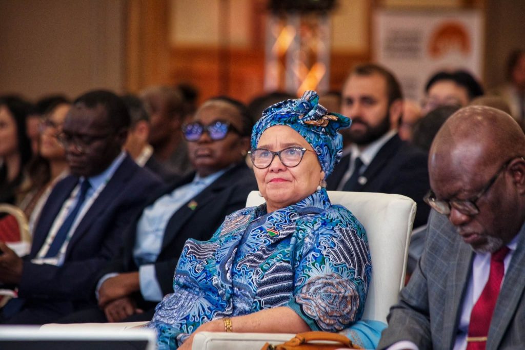 Woman wearing a blue patterned head wrap and glasses sits in a conference audience.