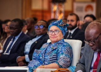 Woman wearing a blue patterned head wrap and glasses sits in a conference audience.