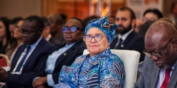 Woman wearing a blue patterned head wrap and glasses sits in a conference audience.