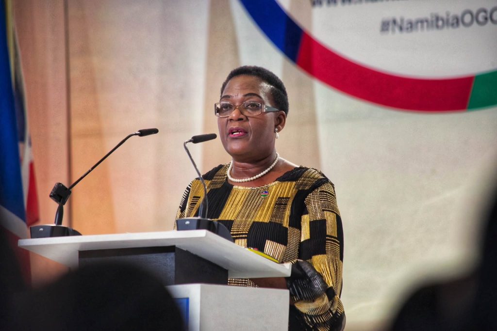 KORNELIA SHILUNGA3 | Mining and Energy Namibia | Namibia’s Leading Mining & Energy News Woman in patterned dress giving a speech at a podium with two microphones and flags in the background.