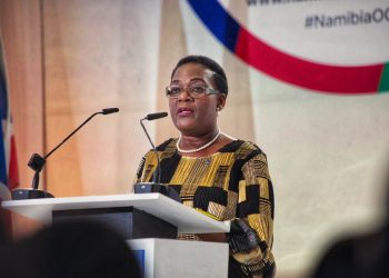 Woman in patterned dress giving a speech at a podium with two microphones and flags in the background.