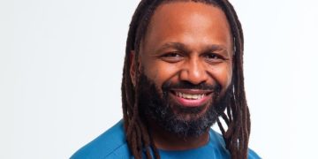 Smiling man with long dreadlocks and beard wearing a blue shirt, facing the camera against a light background.