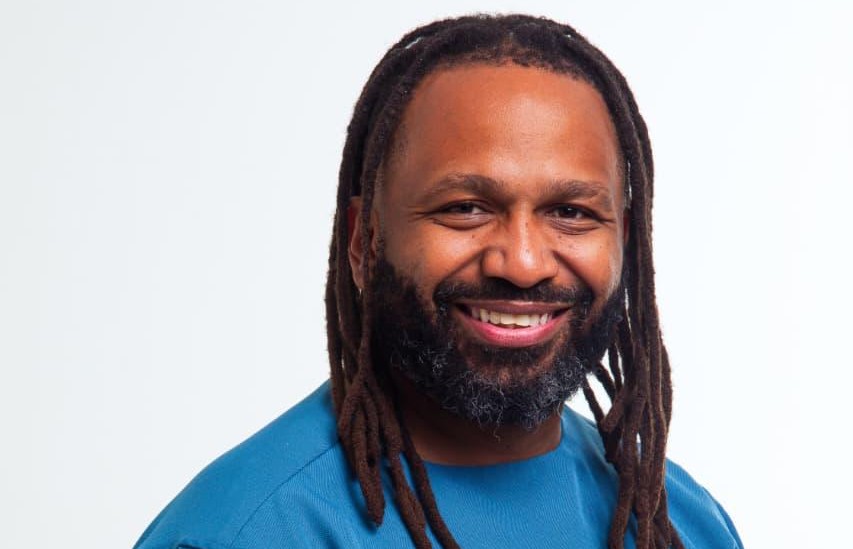 Smiling man with long dreadlocks and beard wearing a blue shirt, facing the camera against a light background.