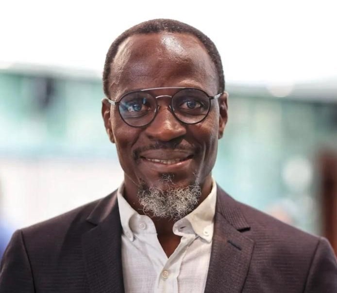 Fillemon Wise | Mining and Energy Namibia | Namibia’s Leading Mining & Energy News Smiling man wearing round glasses, a white shirt, and a dark blazer in a professional setting.