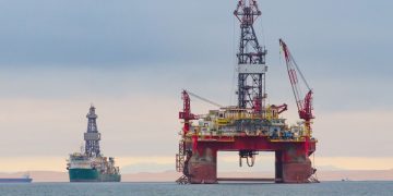 Large offshore oil platform with a tall derrick and cranes in calm sea; a second platform and a ship visible in the distance.