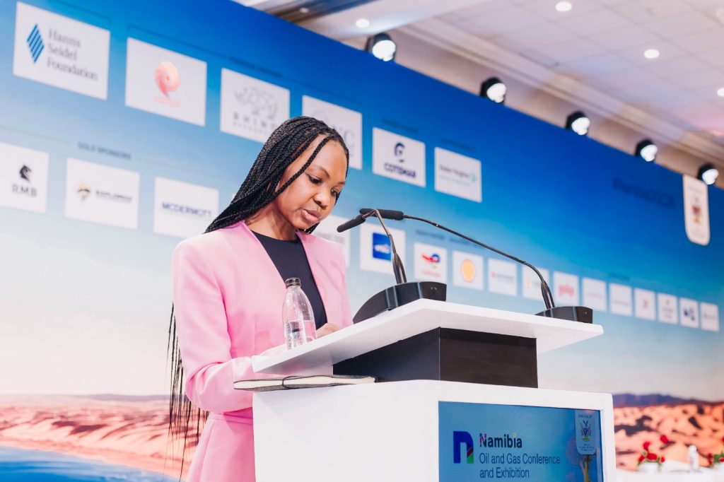 nog2 | Mining and Energy Namibia | Namibia’s Leading Mining & Energy News Woman in a pink blazer speaks at a podium during a conference, with a blue sponsor backdrop behind her.