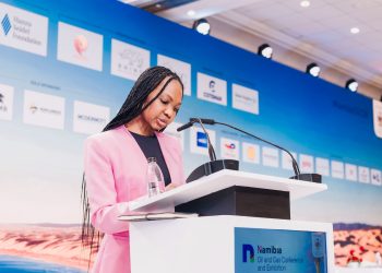 Woman in a pink blazer speaks at a podium during a conference, with a blue sponsor backdrop behind her.