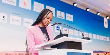 nog2 | Mining and Energy Namibia | Namibia’s Leading Mining & Energy News Woman in a pink blazer speaks at a podium during a conference, with a blue sponsor backdrop behind her.