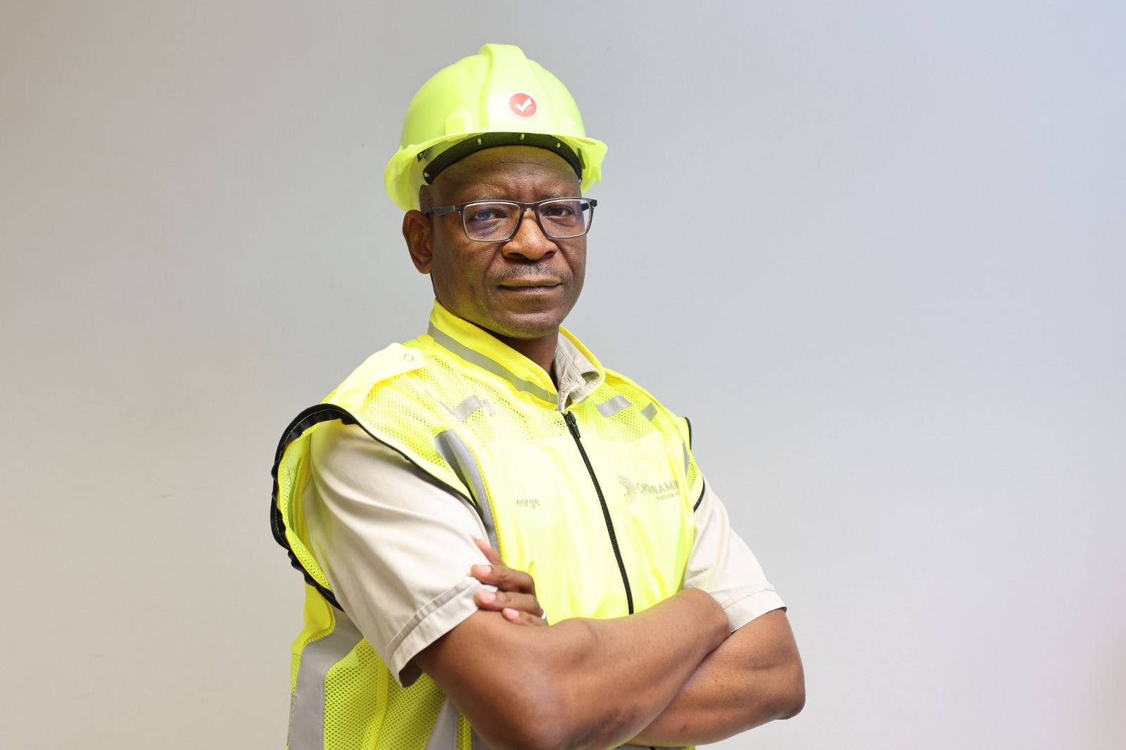 Construction worker wearing a neon yellow safety helmet and high-visibility vest with arms crossed in front of a neutral background.
