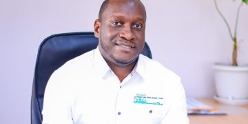 Paulus Mulunga | Mining and Energy Namibia | Namibia’s Leading Mining & Energy News Portrait of a man in a white shirt sitting at a desk, smiling at the camera in an office setting with a plant in the background.