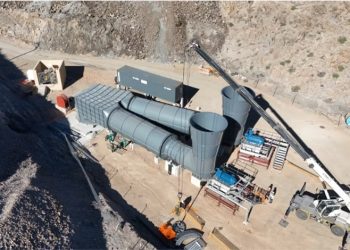 Industrial construction site with large gray cylindrical ducts and a crane lifting material on a dirt lot.