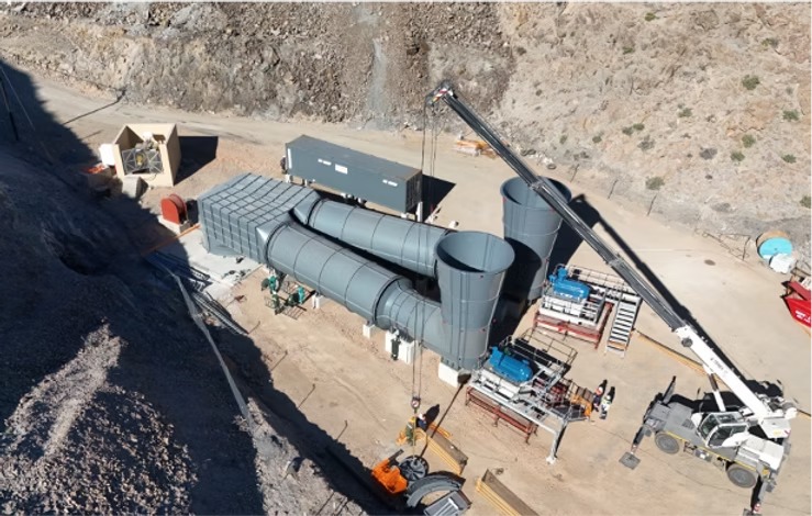 Industrial construction site with large gray cylindrical ducts and a crane lifting material on a dirt lot.