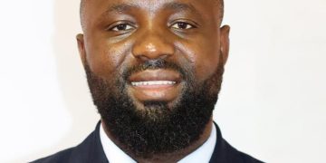 Portrait of a smiling Black man in a navy suit and yellow tie against a light background, a professional headshot.