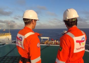 TOTALENERGIES 2 | Mining and Energy Namibia | Namibia’s Leading Mining & Energy News Two workers in orange safety suits and white hard hats stand on an offshore deck, looking out at the open sea toward a distant vessel.