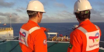 TOTALENERGIES 2 | Mining and Energy Namibia | Namibia’s Leading Mining & Energy News Two workers in orange safety suits and white hard hats stand on an offshore deck, looking out at the open sea toward a distant vessel.