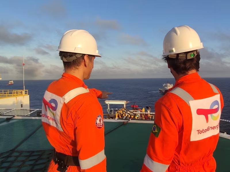 TOTALENERGIES 2 | Mining and Energy Namibia | Namibia’s Leading Mining & Energy News Two workers in orange safety suits and white hard hats stand on an offshore deck, looking out at the open sea toward a distant vessel.