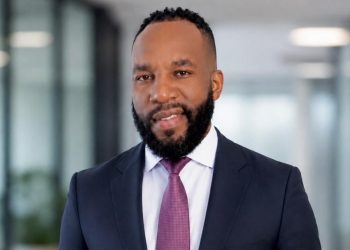 Shiwana Ndeunyema1 | Mining and Energy Namibia | Namibia’s Leading Mining & Energy News Portrait of a smiling Black man in a navy suit and pink tie, standing in a bright modern office corridor.