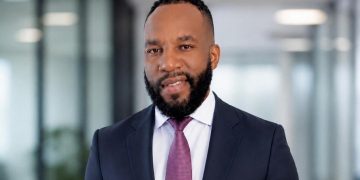 Portrait of a smiling Black man in a navy suit and pink tie, standing in a bright modern office corridor.
