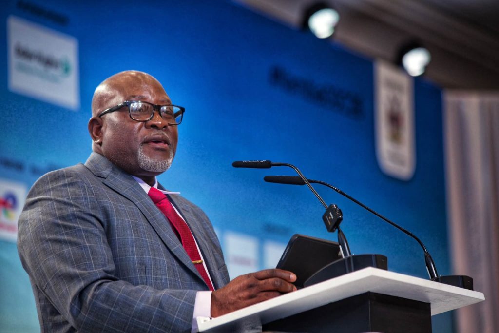 MODESTUS AMUTSE5 | Mining and Energy Namibia | Namibia’s Leading Mining & Energy News A smiling African American man in a gray checkered suit and red tie speaks at a podium with a microphone against a blue backdrop.