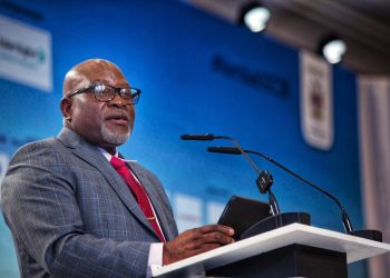 A smiling African American man in a gray checkered suit and red tie speaks at a podium with a microphone against a blue backdrop.