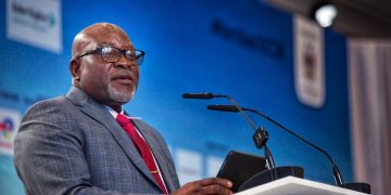 MODESTUS AMUTSE5 | Mining and Energy Namibia | Namibia’s Leading Mining & Energy News A smiling African American man in a gray checkered suit and red tie speaks at a podium with a microphone against a blue backdrop.