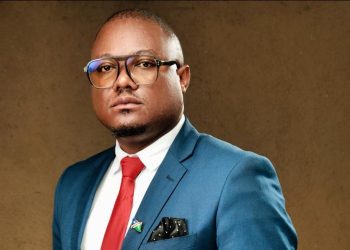 Filleppuss George Ampweya | Mining and Energy Namibia | Namibia’s Leading Mining & Energy News Portrait of a man in a blue suit, white shirt, red tie, wearing glasses, against a brown backdrop.
