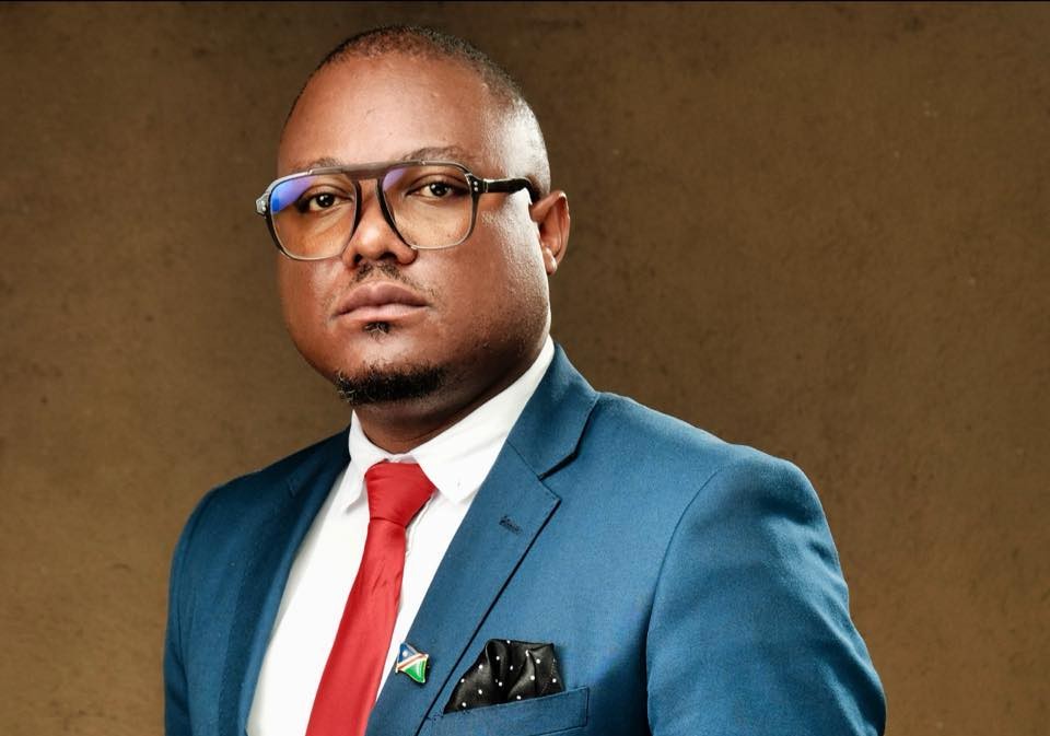 Filleppuss George Ampweya | Mining and Energy Namibia | Namibia’s Leading Mining & Energy News Portrait of a man in a blue suit, white shirt, red tie, wearing glasses, against a brown backdrop.