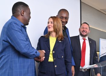 Group of four professionals on a stage, two men shaking hands while a woman in a navy blazer and yellow top looks on, others smiling in background.