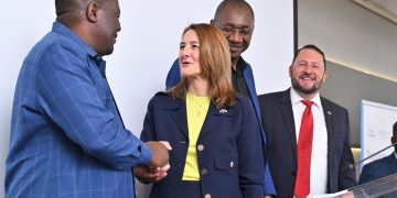 Group of four professionals on a stage, two men shaking hands while a woman in a navy blazer and yellow top looks on, others smiling in background.