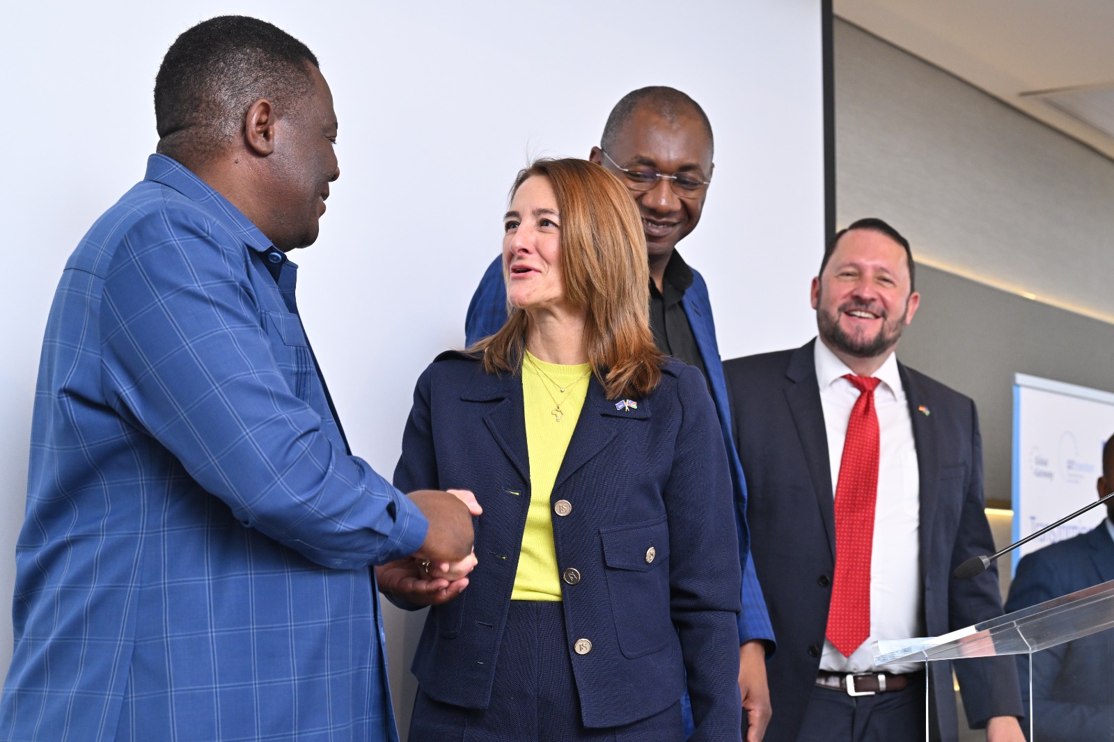Group of four professionals on a stage, two men shaking hands while a woman in a navy blazer and yellow top looks on, others smiling in background.